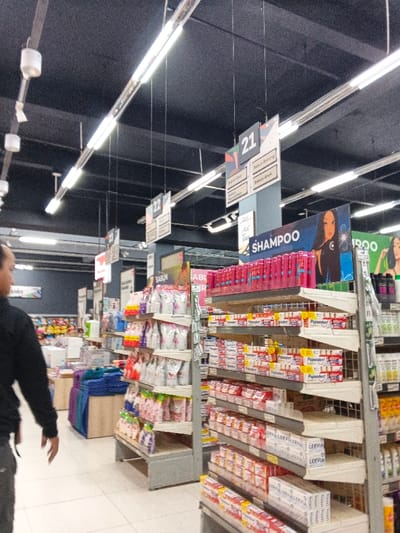 The image captures a supermarket aisle with shelves stocked with products. Signage indicates sections for "SHAMPOO," "SABUN" (soap), and numbered aisle markers "21" and "22". Various brands of shampoo, toothpaste (Pepsodent), and soap (LERVIA) are visible. A person's torso is seen on the left side of the image. The aisle is brightly lit with overhead fluorescent lights.