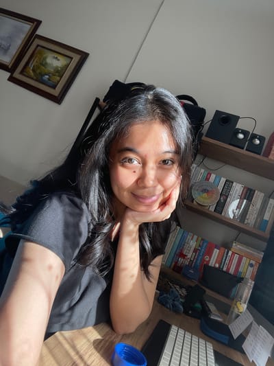 A young woman with dark hair and brown eyes is pictured from the chest up, resting her chin on her hand. She is wearing a black t-shirt. The background consists of a white wall with two framed paintings, a bookshelf filled with books, and a desk with a keyboard and various objects. The scene is illuminated by natural sunlight, creating shadows across the desk and the woman's arm. The location appears to be an indoor residential space, possibly a home office. No elements suggest a specific city or country, let alone Başakşehir, Turkey.