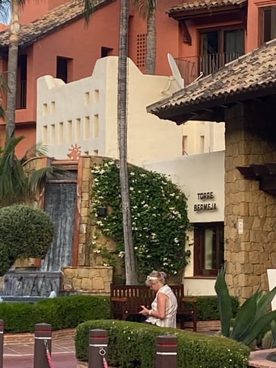 The image depicts the exterior of the Torre Bermeja development in Estepona, Spain. A woman is seated on a wooden bench in the foreground, looking at a red object in her hands. She is wearing a striped sleeveless shirt and a white headband. Behind her, a lush green vine with white flowers climbs a wall, adjacent to a cascading waterfall feature. The architecture is characteristic of the region, with terracotta tile roofs and warm-toned stucco walls. The scene is set in an outdoor area, likely a courtyard or garden, with manicured hedges and palm trees visible.