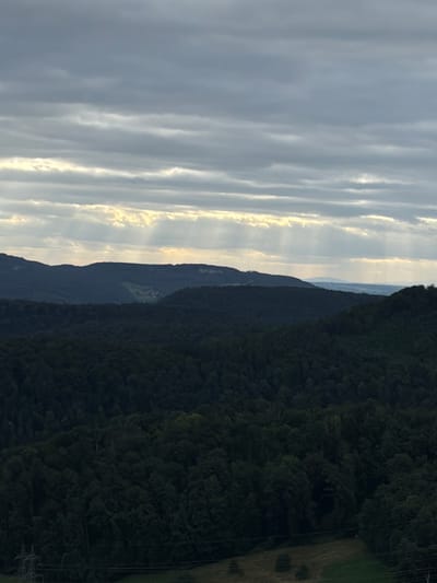 The image shows a landscape view from a high vantage point, likely in or around Sissach, Switzerland. The scene is dominated by rolling hills covered in dark green forests. In the distance, layers of hills recede into the background, with the furthest ones appearing hazy and a lighter blue, suggesting a broad valley or plain. The sky is overcast with dramatic clouds, through which shafts of sunlight are breaking, creating visible light rays. The light rays are most prominent in the upper portion of the image, illuminating patches of the sky and the upper slopes of the hills. In the foreground, a power pylon and some electrical lines are visible on the left side, indicating human infrastructure present in the otherwise natural landscape. The overall atmosphere is moody and somewhat dramatic due to the interplay of dark shadows and bright light. There are no people or animals visible in the frame.