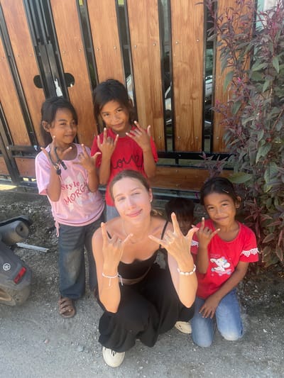In Kertasari, Indonesia, three young girls and a woman are posing for a photograph outside a wooden fence. The girl on the left wears a pink t-shirt and blue jeans, holding a beaded necklace with keys. The girl in the middle wears a red t-shirt. The girl on the right wears a red t-shirt with a cartoon elephant print and blue jeans. A young boy is partially visible behind the woman. The woman, in the foreground, is wearing a black top and black pants. All four individuals in the foreground are making a "rock on" hand gesture. A scooter is parked to the left of the frame, and a bush with red and green leaves is to the right. The ground is covered in gravel.