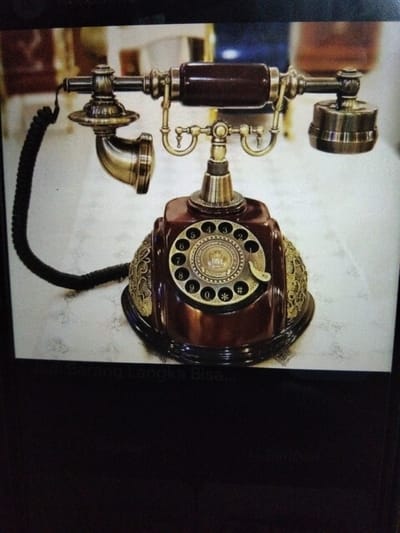 An antique rotary dial telephone with a dark brown body and brass accents sits on a white, textured surface. The telephone has a coiled black cord connecting the receiver to the base. The receiver itself is brass and has a dark brown handle. A circular dial with numbers zero through nine, as well as asterisk and hash symbols, is prominently featured on the front of the base. Decorative brass filigree adorns the sides of the base. The background is blurred but suggests an indoor setting with some architectural elements. The image has a watermark at the bottom that reads "Barang Langka Bisa". The location in the city of Kualaraja, Indonesia is not explicitly indicated by any visual cues in the image.