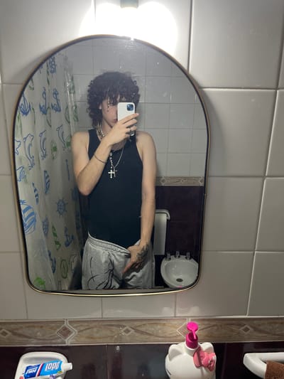 A mirror selfie, presumably taken in a bathroom, shows a young person with curly dark hair. They are wearing a black tank top and gray sweatpants. They are also wearing multiple necklaces, including one with a cross. The person is holding a white smartphone, presumably to take the photo. The background includes a patterned shower curtain, white tiled walls, a sink, and various bathroom items, including a Hello Kitty-themed soap dispenser and a tube of toothpaste. There is no specific information available to indicate a location in Bahía Blanca, Argentina.