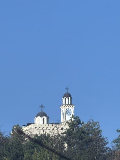 The image shows a close-up view of a white stone building with a dark dome topped by a cross, and a white clock tower with a cross on top, both situated on a hill. The building has arched windows and a crenelated wall. The clock face shows the time as approximately 10:10. The scene is set against a clear blue sky, with the lower part of the image dominated by a dense growth of green trees. The overall impression is of a historic or religious structure overlooking a natural landscape. This is the Hadzhidimovo Clock Tower in Hadzhidimovo, Bulgaria.