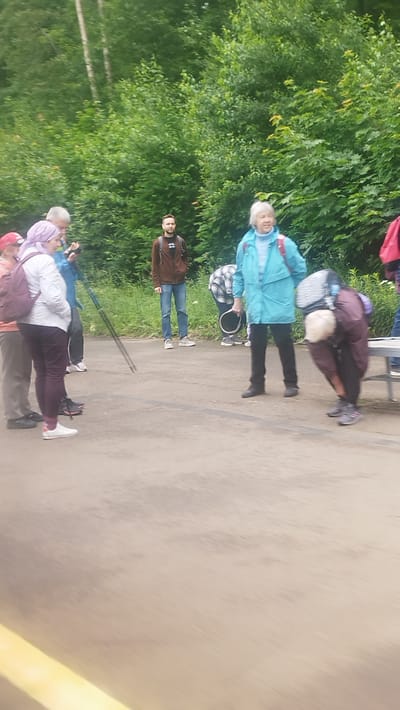 This media file shows a group of people gathered outdoors in what appears to be a park or wooded area. The setting is characterized by lush green trees in the background, suggesting a daytime scene in a temperate climate, likely during warmer months. The ground is a paved or asphalt surface.
Several individuals are visible, appearing to be engaged in a casual gathering or possibly preparing for an outdoor activity. In the foreground, a person wearing a light purple headscarf and a white top with dark pants is facing away from the camera, seemingly interacting with someone to their left. To their left, another individual with gray hair and a blue jacket is partially visible.
Further back, a man in a brown jacket and jeans stands looking towards the group. To his right, a person in a teal-colored jacket is facing the camera, with a backpack on. Another individual is bent over, possibly tending to something on the ground or adjusting equipment.
Objects present include backpacks worn by some individuals, and a pair of walking poles held by someone in the center. The overall mood appears relaxed and social. There are no visible signs or text in the image. Given the location context of Toksovo, Russia, the scene likely depicts a recreational outing in a natural setting. The lighting suggests it is daytime, and the greenery indicates a season with active vegetation.