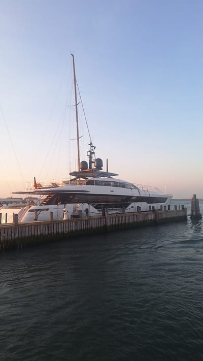 A large, white motor yacht with a prominent mast is docked at a wooden pier in the waters of Nantucket, Massachusetts. The yacht is sleek and modern, featuring multiple levels and dark accents. The sky above is clear and pale blue, transitioning to a soft pink near the horizon, indicating either sunrise or sunset. The water is dark and rippled. Several wooden pilings are visible along the pier, and a single, weathered piling stands in the water to the right of the yacht. The overall scene is serene, depicting the yacht moored in a harbor environment.