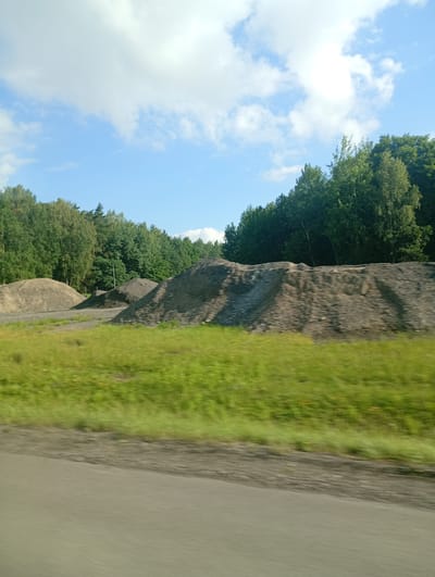 The image shows a landscape with large piles of gravel and sand in the foreground, a line of trees in the background, and a blue sky with clouds above. The ground in the foreground appears to be a dirt road or path. The lighting suggests it is daytime. Based on the provided hints, the location is identified as Mysłowice, Poland.