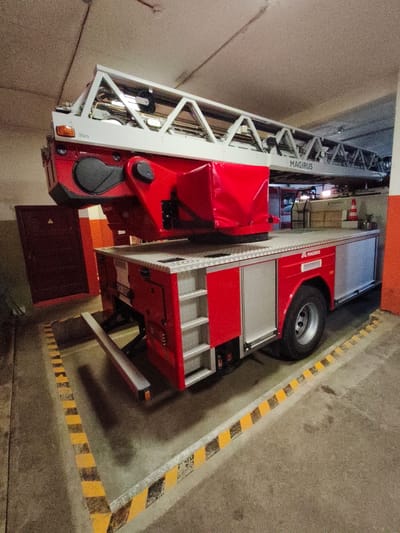 A red and silver Magirus fire truck with an extended ladder is parked indoors, likely in a fire station or garage. The ladder extends upwards and across the frame. The truck has "MAGIRUS" branding visible on the side and on the ladder mechanism. The environment appears to be a concrete floor with yellow and black striped safety markings. In the background, there is a closed dark red door and an orange and white striped wall. The scene is well-lit.