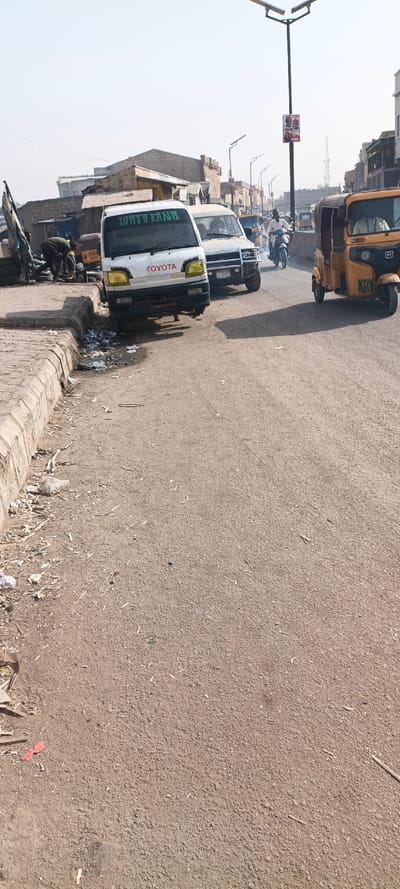 This image captures a daytime street scene in Kaura Goje, Nigeria. The foreground presents a dusty, paved road with a raised concrete curb on the left, bordering an unpaved shoulder scattered with litter, plastic bags, and wood debris.
On the left side of the frame, a person in dark clothing is bent over near a collection of dark objects, possibly tires, on the shoulder. Adjacent to this, a white Toyota minivan with "WATH RANA" written in green on its windshield and yellow-tinted headlights is positioned with its front wheels on the unpaved shoulder and its rear on the paved road; its front bumper appears to be missing.
Behind the Toyota minivan, a second white van equipped with a large metal front bumper guard is visible. Further down the road, an individual on a motorcycle, wearing a light-colored shirt, rides toward the right side of the frame, with visible exhaust fumes trailing. To the far right, a yellow auto-rickshaw, marked "KRY" on its side, is in motion, heading away from the viewer. Other indistinct vehicles, including additional auto-rickshaws, and figures are present further down the street.
The background reveals a series of single and multi-story buildings lining both sides of the street, some appearing to be constructed with exposed brick or concrete, and some in various states of completion or disrepair. Modern streetlights with solar panels are erected along the thoroughfare, one of which displays a multi-person political poster. The sky is a uniform, light, hazy blue.