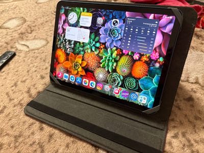 This media file shows a close-up, slightly overhead view of a tablet device resting on a textured, beige surface, possibly a rug or blanket. The tablet is open and positioned at an angle, showcasing its screen. The screen displays a vibrant wallpaper featuring a colorful arrangement of succulents.
Several widgets are active on the tablet's home screen. On the left, there's a clock widget showing an analog time of approximately 10:10, and a calendar widget displaying "ПЯТНИЦА 26" (Friday 26) with a note "Сегодня событий нет" (No events today). Below these, there's a calendar for June. To the right, a weather widget for "Засечное" (Zasechnoye) indicates a temperature of 11°C and mentions "В основном облачно" (Mostly cloudy) with a forecast range of 10°C to 21°C. Further right, another weather forecast displays temperatures for the next few days.
App icons are visible in a row at the bottom of the screen, including Camera, Settings (with a notification badge), and others like an email icon, a music app, a shopping app labeled "Ozon," and a calendar icon showing "26". The top right corner of the screen shows a battery indicator at 66% and the time "00:39 Пт 26 июня" (00:39 Friday June 26).
A dark grey tablet case is partially visible, holding the device. To the left of the tablet, a black remote control lies on the textured surface. The overall lighting suggests it's daytime, but the screen brightness is high. The scene appears to be indoors, with no distinct location cues beyond the "Zasechnoye" text on the weather widget.