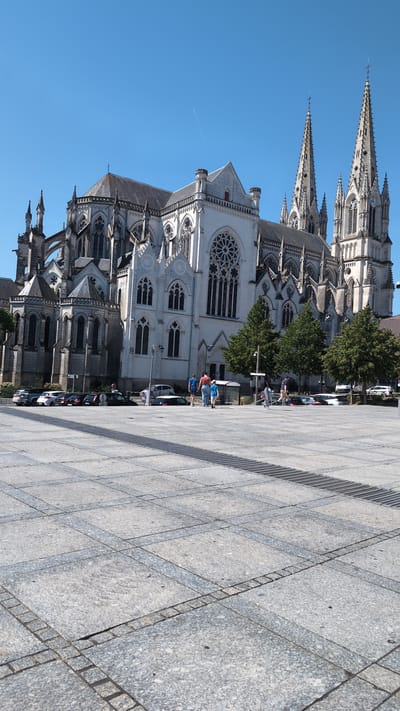 A wide, sunlit shot captures the exterior of the Cathédrale Saint-Louis in Cholet, France, under a clear blue sky. The imposing gothic cathedral dominates the left and central portions of the frame, its white stone facade adorned with intricate architectural details, including pointed arches, spires, and a large rose window. To the right, two tall, slender spires rise prominently, one featuring a clock face.
In the foreground, a vast, paved plaza stretches across the bottom half of the image. The ground is laid with large, light grey stone tiles, some of which show subtle variations in color and texture, accented by darker grey borders forming a grid pattern. A dark metal grate runs diagonally across the plaza from the mid-ground towards the lower right corner.
A few individuals are visible in the mid-ground, walking on the plaza. Three people, two adults and a child, are walking away from the camera towards the cathedral entrance. Further to the right, another person is walking in the same direction. Several parked cars are visible along the edge of the plaza near the cathedral. Lush green trees are scattered along the base of the cathedral, providing a contrast to the stone architecture. The overall scene depicts a serene urban environment on a bright, clear day.