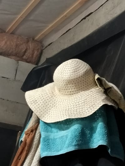 The image shows a white straw hat with a wide brim, draped over what appears to be a teal-colored towel. Behind the hat and towel, there is a dark fabric, possibly a coat or a large bag, and further behind that, a brick wall. The ceiling above is unfinished, with exposed wooden beams and insulation material visible. The scene appears to be indoors, in an area that is under construction or renovation, or possibly a storage area. There are no people visible in the image. The lighting is dim. The location within the city of Kudashivka, Ukraine, is not indicated by any visual cues in the photograph.