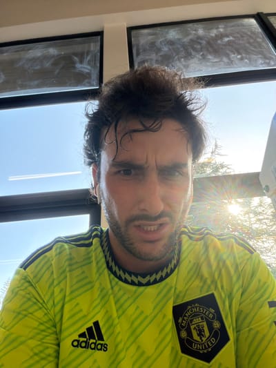 A man wearing a yellow Manchester United jersey takes a selfie. His dark hair is slightly messy, and he has stubble. The bright sun streams through a window behind him. The Adidas logo is visible on his sleeve, along with the Manchester United badge on his chest. The background includes a window with grimy panes. He is looking at the camera.