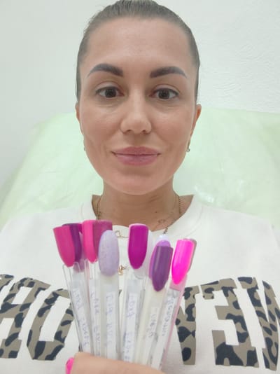 A woman is holding several nail polish swatches in front of her chest, facing the camera. She has dark hair pulled back and is wearing a white sweatshirt with a leopard print design. The background appears to be a white wall with a light green draped fabric. The lighting is bright and even, suggesting an indoor setting. There is no identifiable information about the location in Komsomolsk-on-Amur, Russia.