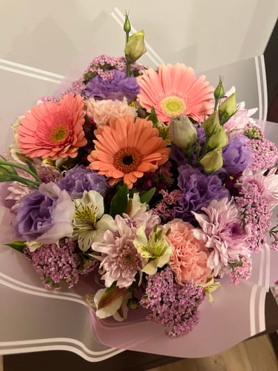 This media file is a close-up, top-down view of a vibrant bouquet of flowers. The arrangement features a variety of blossoms in shades of pink, orange, purple, and white, with accents of green foliage. Prominent flowers include large, round gerbera daisies in pink and orange, delicate pink carnations, and ruffled purple lisianthus. Smaller, clustered flowers in pink and purple add texture and depth to the bouquet. Some flowers are fully bloomed, while others appear as buds, suggesting the bouquet is fresh.
The flowers are artfully arranged and partially wrapped in a soft, lavender-colored paper that has subtle white stripes. The background is out of focus, showing a neutral gray surface with the same white stripe pattern, suggesting the bouquet might be resting on a table or surface with decorative wrapping. A sliver of a darker, possibly wooden surface, is visible on the right side of the frame.
There is no discernible activity or event taking place beyond the display of the bouquet itself. The lighting appears to be even and diffused, possibly from indoor lighting, and there are no indicators of the time of day or weather conditions. No text is visible in the image. The overall impression is one of beauty, freshness, and a celebratory or decorative purpose, common for floral arrangements in many settings, including events or as a gift.