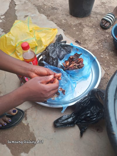 In Dokan Tofa, Nigeria, a close-up shot captures a person's hands processing dried chilies, positioned on a concrete ground with visible cracks and patches of dampness. The hands, with dark skin and dark purple nail polish on the toes of a visible foot in a sandal, are actively separating chilies from their stems. They are working over a large, circular, metallic tray lined with blue plastic bags. Within the tray, several dried chilies are arranged, some partially open and revealing seeds. Adjacent to the tray, a clear glass bottle with a red cap, containing a golden liquid, lies on its side next to a crumpled yellow plastic bag. A black plastic bag, also crumpled, is situated next to the tray. In the background, a large black bucket and a blue plastic bowl are partially visible. The overall environment suggests an outdoor setting, possibly a market or a domestic workspace.