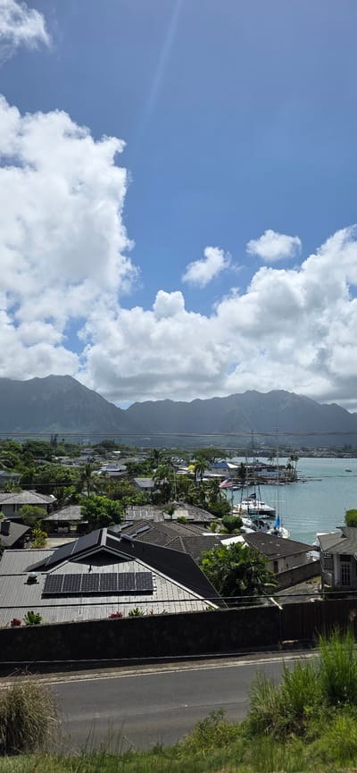 This is a high-angle, vertical, outdoor, daytime photograph overlooking a residential area in Kaneohe, Hawaii, United States of America. The scene captures a suburban neighborhood with a backdrop of lush green mountains and a calm blue body of water. In the foreground, a road runs horizontally across the bottom of the frame, with grassy areas and vegetation on either side.
Dominating the middle ground are the rooftops of houses. Several houses feature dark tiled roofs, and one prominent house in the lower left has a dark metal roof covered with an array of solar panels. Another house to the right has a lighter-colored roof. Beyond these immediate homes, more houses with varying roof colors and styles are visible, interspersed with dense green trees and palm trees.
The water in the middle right of the image is a tranquil bay or harbor, dotted with several sailboats and yachts. An American flag is visible on a flagpole at the water's edge. The water appears calm and reflects the sky.
In the background, a majestic mountain range, likely the Koʻolau Range, forms a dramatic silhouette against the sky. The mountains are covered in dense vegetation and are partially obscured by clouds, particularly towards the peaks.
The sky is a vibrant blue, with numerous fluffy white cumulus clouds scattered across it. A faint sun glare or lens flare streaks diagonally from the upper left towards the center of the frame. There are no people or animals visible in the photograph. The overall impression is a serene and picturesque coastal residential landscape.