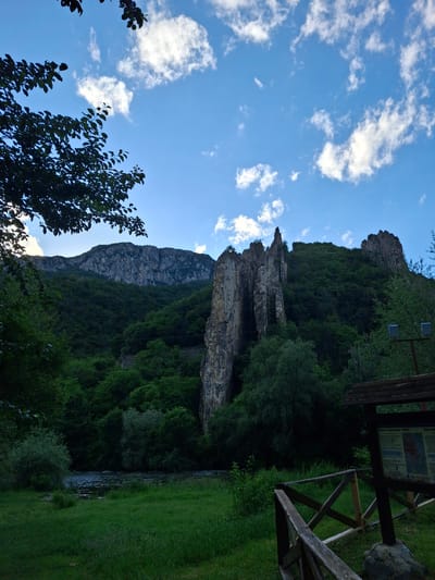 This image captures a vibrant natural landscape under a bright blue sky with scattered white clouds. The primary subject is a towering, rugged rocky cliff face, partially covered by dense green trees and vegetation. Several thin cables or lines are visible descending the cliff, possibly for adventure activities or structural support. To the right, a wooden structure, likely a viewing platform or picnic area, holds an interpretive sign with text in Cyrillic. Notable legible text on the sign includes "Римска крепост" (Roman fortress), "Вратцата" (Vratsata), and "Лютиброд" (Lyutibrod), confirming the location in Lyutibrod, Bulgaria. Two square floodlights are mounted on a pole near the top right, above the cliff. At the bottom right, a calm river or stream flows, flanked by lush green grass and more trees, reflecting the sky. The time of day appears to be bright daylight, possibly morning or afternoon, with clear weather. No people or explicit activities are visible, but the man-made structures suggest a visitor-friendly natural attraction.