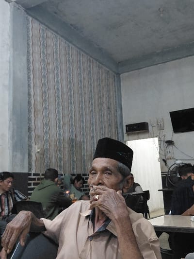 An elderly male, dressed in a light brown collared shirt and a black songkok with green embroidery, is prominently featured in the foreground, seated within an indoor establishment in Mns Alue, Indonesia. He holds a lit cigarette to his mouth with his right hand, displaying a gold-colored ring on his left ring finger. His face and hands show visible signs of age. In the background, multiple individuals are seated at dark tables; one male in a striped shirt is visible to the far left, and another in a green hoodie is in the mid-background. The interior features a patterned wallpaper with vertical wavy stripes in beige and light blue tones on the left wall, with a darker tiled section below it. The right wall is plain light-colored, and the ceiling is unfinished grey concrete with visible beams. Exposed electrical wiring, a black speaker, and a wall-mounted dark screen are visible on the right wall, alongside a partial view of a fan.