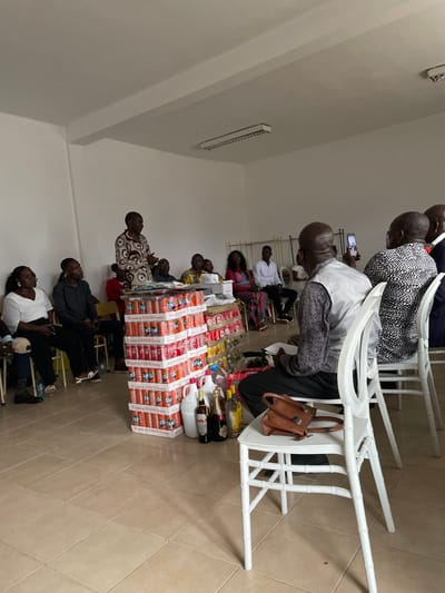 This image captures an indoor gathering in what appears to be a community hall or event space in Luanda, Angola. The room features light-colored walls and a tiled floor.
Numerous people, predominantly adults, are seated on white plastic chairs arranged in rows, facing towards the right side of the frame. The perspective is from the front-left of the assembled group. In the foreground and middle ground, individuals are seen observing or engaged with their personal items, such as mobile phones. One person in a light-colored shirt and dark trousers has a brown leather handbag hanging from the back of their chair. Another individual nearby appears to be holding papers.
The central focus of the scene is a substantial display of various beverages arranged on what seems to be a table or platform. The base of this display is formed by stacked boxes of soft drinks, with "FANTA" and "Coca-Cola" branding prominently visible on the cans and cartons. The text "THE COCA-COLA COMPANY" is also legible on some boxes. On top of these stacked soft drink cases, an assortment of bottled alcoholic beverages, including what appear to be whiskey and wine, are meticulously arranged alongside several clear plastic jugs.
The event suggests a form of presentation, perhaps a ceremony involving gifts or donations, or a social gathering where these items are displayed for distribution. The lighting within the room is artificial, with a fluorescent light fixture visible on the upper part of the left wall. The attendees are dressed in a mix of casual and semi-formal attire, and their expressions appear calm and attentive.