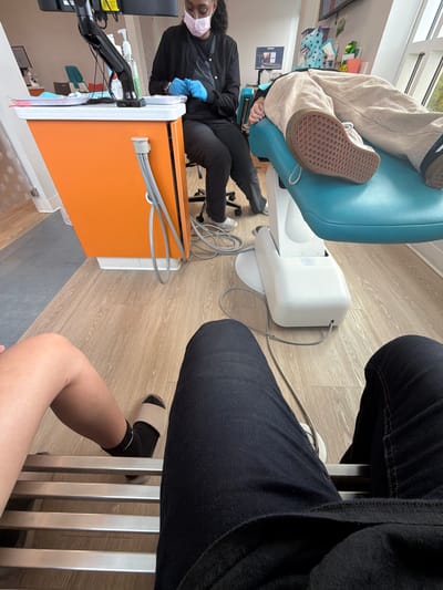 The image shows a top-down view of a dental office or examination room, oriented sideways. Two individuals are partially visible, along with medical equipment.
On the left, a person wearing a black scrub top, blue gloves, and a pink face mask is seated and appears to be working with their hands. They are positioned near an orange and white cabinet-like piece of equipment, which has medical tubing connected to it. Another person, whose legs are prominently featured, is seated in the foreground wearing dark blue jeans and slides on their feet. Their legs are crossed, and they are leaning against a metal chair base.
The setting is indoors, with a light-colored wood-grain floor. In the background, a teal dental chair is visible, along with a white piece of equipment attached to it. There are also shelves with various items, including what looks like a blanket or throw with a polka dot pattern, and some books or folders. The overall impression is a clinical or professional environment. The lighting suggests it is daytime, and there are no clear indications of weather conditions. No specific emotions are evident from the visible parts of the subjects. The visible text includes labels on the equipment, but they are not legible in this image. The location is identified as Chesapeake, USA.