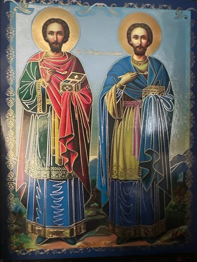This is an image of an icon depicting two male figures with halos. Both figures have dark hair and beards, and are dressed in robes. The figure on the left wears a green robe with a red garment draped over it, and holds a golden box with a cross symbol. The figure on the right wears a blue robe over a yellow garment and also holds a golden box with a cross symbol. The background is a textured blue with decorative borders. The image appears to be a religious icon, likely displayed indoors. There is no discernible indication of location within the city of Marten, Bulgaria, nor any details about the environment or context that would suggest such a location.
