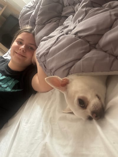 This image shows a young person lying in bed, with their head resting on their hand. A small white dog is peeking out from under the covers next to them. The room is lit by artificial light, and a shelf unit with a television is visible in the background. The scene appears to be indoors, possibly a bedroom. There is no information available to suggest this image is located in Shepetivka, Ukraine.