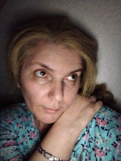 A woman with blonde hair is captured in a close-up, selfie-style photograph, her face tilted slightly upwards and to the right. She is resting her chin on her right hand, which is positioned under her jaw. Her expression appears pensive, with her eyes looking off into the distance. She is wearing a teal-colored blouse adorned with a floral pattern of pink and black flowers. A silver bracelet is visible on her left wrist. The background is a textured wall, dimly lit, with shadows creating a somber atmosphere. The scene is indoors, with the lighting suggesting it could be nighttime or a dimly lit room. No specific location in Al Mas'oudi, United Arab Emirates can be determined from the visual information provided.