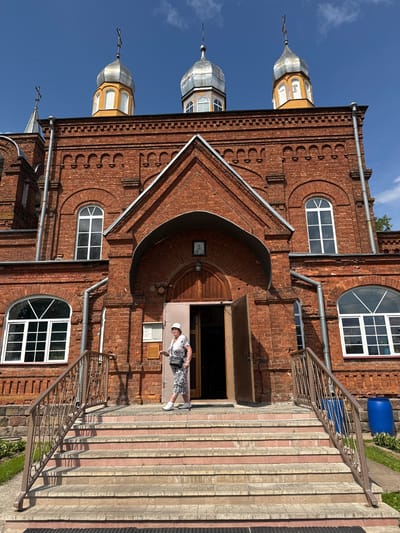 An elderly woman in a white hat and a patterned dress stands on the steps of a red brick church in Chudovo, Russia. She holds a walking stick in her right hand. The church features arched windows, a prominent entrance with a wooden door, and three onion domes topped with crosses against a clear blue sky. A metal railing lines the stairs, and two large blue barrels are visible to the right of the church. The scene is illuminated by bright sunlight, casting shadows on the steps.