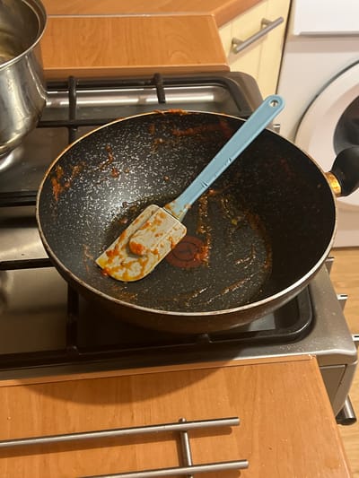 A close-up, eye-level shot shows a dirty frying pan with a spatula inside, resting on a gas stovetop. The pan has remnants of a red sauce splattered around its interior. The spatula, made of light blue plastic and white silicone, is angled across the pan, also coated with the sauce. To the left of the pan, a stainless steel pot with a clear liquid is partially visible. The stovetop is black and appears to be part of a kitchen environment with wooden cabinets and a portion of a white appliance in the background. The overall scene suggests a recently used cooking surface. The location in Skawina, Poland is not determinable from the image.