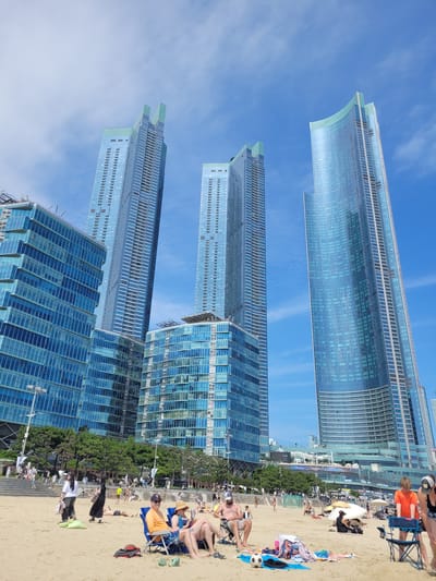 This is an eye-level, medium shot photograph taken on a sunny day at Haeundae Beach in Busan, South Korea. The foreground is dominated by a sandy beach with several people relaxing on lounge chairs. A man in an orange shirt and a woman in a blue bathing suit are seated in blue chairs, with beach towels and a soccer ball on the sand in front of them. Other beachgoers are scattered across the sand, some lying down, others standing and interacting.

In the mid-ground, lush green trees line the edge of the beach, providing a transition to the urban landscape. Behind the trees, a complex of modern, glass-facaded skyscrapers rises dramatically against a bright blue sky with scattered white clouds. These buildings are a prominent feature of the Haeundae district, known for its high-rise apartments and hotels. One particularly distinctive building in the background has a curved, shimmering glass exterior, reflecting the sunlight. The architectural style is contemporary, characterized by sleek lines and expansive glass panels. The overall scene depicts a vibrant urban beach environment, with people enjoying leisure activities against a backdrop of impressive modern architecture.