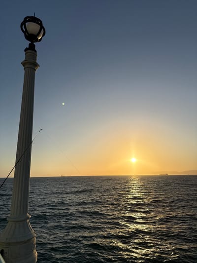 This image depicts a sunset over the Pacific Ocean, viewed from the Manhattan Beach Pier in Manhattan Beach, California, United States of America. In the foreground, a white lamppost with a decorative base stands to the left, its shadow cast towards the right. A fishing rod is propped against the lamppost, with its line extending towards the ocean. The sun is setting on the horizon, casting a warm golden glow across the water and the sky. The ocean is relatively calm with gentle waves, and several ships are visible in the distance. The sky transitions from a clear blue at the top to a soft orange hue near the horizon.