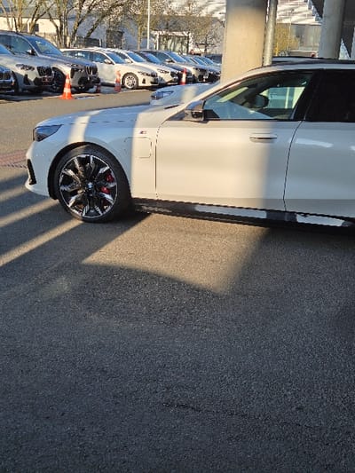 The scene depicts an outdoor vehicle display area at a BMW dealership in Sprendlingen, Germany. In the foreground, the front-right section of a white BMW i4 M50 is prominently positioned on asphalt pavement, showcasing black multi-spoke wheels with red brake calipers and an M badge on the front fender. Direct sunlight illuminates portions of this vehicle, casting defined shadows onto the pavement. In the midground, a linear arrangement of multiple BMW vehicles, primarily SUVs and sedans in light grey and white finishes, are parked. Two orange traffic cones are visible among these parked vehicles. The background features a modern, multi-story commercial building with a structured facade, alongside several bare deciduous trees. A substantial light-colored concrete support pillar stands on the right side of the frame. No individuals are present, and no actions are occurring within the static scene.