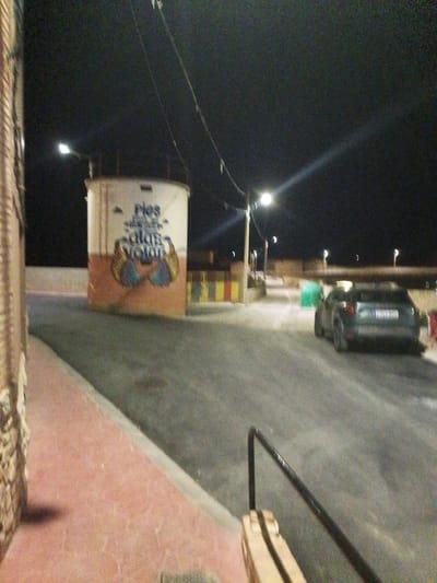 At night, on a street in Mainar, Spain, a cylindrical structure with faded text and artwork stands to the left of the road. The structure has a white top section with "Pies" and what appears to be stylized wings painted on its lower, orange section. The road is paved and curves to the right. A dark-colored car is parked on the right side of the road, facing away from the camera. Streetlights illuminate the scene, casting long shadows. A brick wall is visible on the far left of the image.