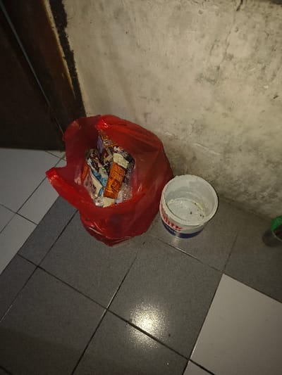 The image depicts an indoor scene in the city of Tarqumiyah, Palestinian Territory. Centrally located is an open red plastic trash bag, filled with various discarded items including food wrappers and general refuse. To the right of the bag, a white plastic bucket with blue accents is visible, appearing to contain some light residue or liquid at its bottom. The objects rest on a tiled floor, composed of dark grey square tiles predominantly in the foreground, transitioning to a section of white square tiles on the far right. The background features a textured, light grey concrete wall, showing signs of wear and minor discoloration. On the left side of the frame, a vertical dark wooden structure, possibly a door frame or partition, is partially visible. No individuals are present, and no actions or interactions are occurring. The environment appears utilitarian or a storage area, illuminated by a light source creating reflections on the tiled floor.