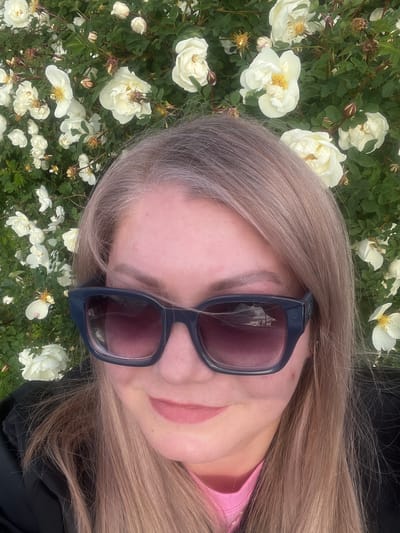 An angled, close-up selfie captures a person with long, light blonde hair, streaked with grey or silver near the roots, partially obscuring their face. The person is wearing dark-framed sunglasses with reflective lenses, beneath which a hint of pink lipstick is visible. A pink garment can be seen near the neck, with a black outer layer visible on the right. The background is a dense bush filled with numerous white, open-faced flowers, likely roses, characterized by their delicate petals and prominent yellow stamens, interspersed with green leaves. Some withered flowers and buds are also present. The scene is brightly lit by natural daylight, suggesting a sunny or overcast day. The composition implies a relaxed pose, possibly lying down or tilting the head to capture the surrounding flora. No visible text is present. The context provided places this scene in Shud'ya, Russia.