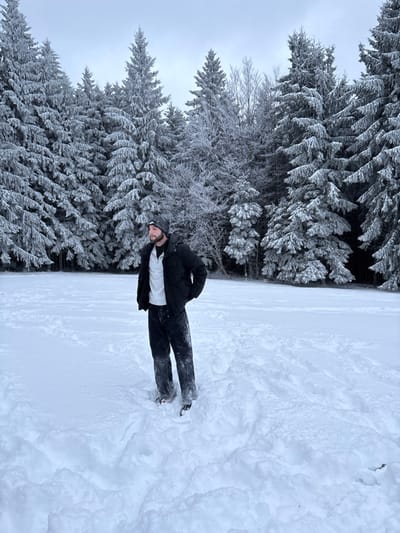 The scene, located in Le Hohwald, France, depicts a male subject standing amidst a snow-covered clearing. He is attired in a dark beanie, a dark winter jacket, a light-colored collared shirt, and dark trousers, with his hands in his jacket pockets. The lower portions of his trousers are visibly damp from contact with the deep snow. The ground is uniformly covered in fresh snow, with depressions evident around the subject's feet. Behind the subject is a dense forest composed primarily of coniferous trees, heavily coated in snow and rime ice. The sky above is overcast and uniformly grey.