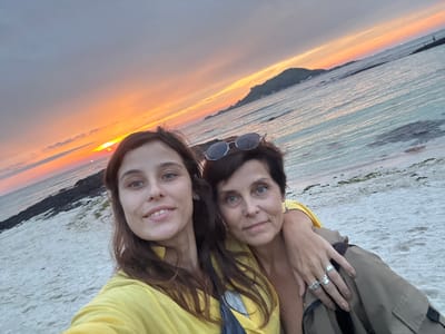This is a portrait of two individuals taken from a low angle. The image is oriented such that the horizon is vertical and the subjects are upside down. The subjects are an adult and a younger person, both with dark hair and fair skin, posing close together. The adult on the left has blue eyes and is wearing silver rings on their left hand, one of which is a band and another appears to have a stone. The younger person on the right has fair skin and is smiling. They are both wearing yellow tops. Behind them, the scene depicts a body of water, possibly the sea, with gentle ripples. The water transitions into a beach or shoreline with some sparse vegetation. In the background, a dark landmass, likely a coastline or island, is visible. The sky displays a vibrant sunset with warm hues of orange, yellow, and pink blending into a softer grey. The sun itself is partially obscured by clouds. The setting suggests an outdoor location, likely a beach or waterfront, during sunset. The mood appears cheerful and relaxed, indicated by the smiles and close proximity of the subjects. The time of day is clearly sunset. There is no visible text in the image. The location context of Jeju, South Korea, aligns with the coastal scenery and the presence of islands in the distance.