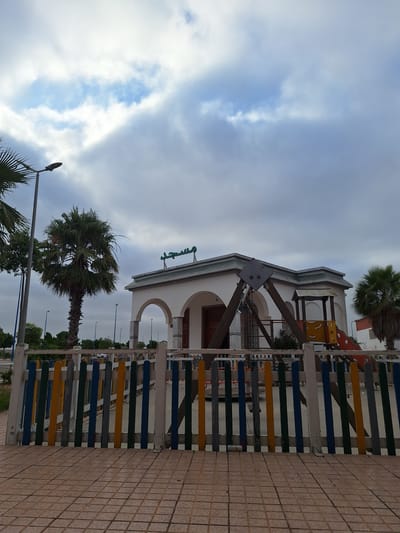 The image depicts a playground area with a small building, a swing set, and a slide in Oulad Hcine, Morocco. The building is white with arched doorways and decorative trim. Arabic text is visible on the roof of the building. A colorful wooden fence separates the playground from a tiled walkway. Palm trees and streetlights are also present. The sky is overcast with scattered clouds. There are no people visible in the image.