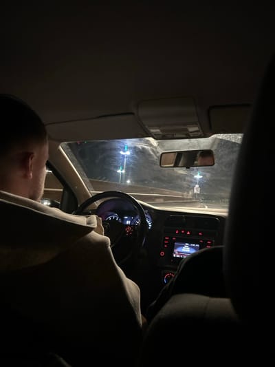 Inside a dimly lit car, a person in a hoodie sits in the driver's seat. The dashboard, steering wheel, and a rearview mirror are visible. The driver's head is turned to the left. Outside, through the windshield, streetlights or other light sources can be seen. The image has a dark and somewhat grainy aesthetic.