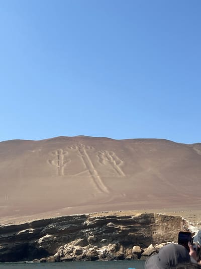 This media file captures a clear daylight scene featuring the ancient Paracas Candelabra geoglyph. The image is oriented vertically but depicts a horizontal landscape.

The primary subject is the massive geoglyph, carved into a light brown, sandy slope that rises from a body of water. The geoglyph, often referred to as the "Candelabra of the Andes," is a large, trident-like figure. It features a prominent central vertical shaft, from which two main upward-curving arms extend outwards, roughly symmetrical. Each of these arms, and the top of the central shaft, terminates in a distinctive three-pronged or petal-like design. The lines of the geoglyph are deeply incised, casting subtle shadows that highlight its form against the sandy surface.

To the right of the sandy slope, a dark, layered rocky cliff face plunges down to the water. The water itself is calm and reflects the light. Above and to the right of the cliff, a few individuals are partially visible, appearing to be on a boat. They are looking towards the geoglyph, with at least one person holding up a mobile phone, likely capturing photos or video of the impressive artwork.

The sky above is a uniform, clear blue, indicating bright and sunny weather. The strong illumination suggests it is mid-day. The scene depicts an activity of tourism and observation of a significant historical landmark, located in the coastal region near Puerto General San Martin, Peru. There is no visible text within the image.