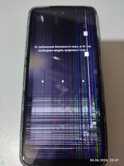 The image shows a close-up of a damaged smartphone screen. The screen displays distorted colorful lines and patterns, indicating a significant malfunction or physical damage. Some white square shapes and circular white dots are also visible on the screen. Text in Russian is partially visible, appearing to be a system message. The top right corner of the screen shows a timestamp: "30.06.2026 22:47". The phone is lying on a white surface, and the edge of the phone's black casing is visible around the screen. The overall impression is that of a broken electronic device, possibly in a home or office setting given the neutral background. The context of Iskitim, Russia, suggests the location where the image was taken or where the phone is located. The time and date indicate a late evening on a summer day in the near future. There are no people visible in the image.