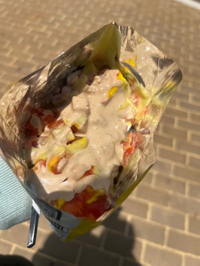 This close-up image shows a person enjoying a casual, outdoor snack in Guayaquil, Ecuador. The main subject is a large serving of food contained within what appears to be a metallic or foil-lined bag, likely a cut-open chip bag, held at the bottom right by a hand wearing a light blue or mint green ribbed sleeve.

The snack itself is a medley of diced ingredients generously coated in a thick, creamy, light brown sauce, possibly a flavored mayonnaise or local dressing. Visible components include vibrant red diced pieces, strongly suggesting tomatoes, and yellow diced elements that could be cooked corn, cheese, or another vegetable. Smaller, lighter-colored diced ingredients, possibly onions or similar, are also mixed throughout. A dark, metallic utensil, likely the handle of a fork or spoon, is visible protruding from the right side of the bag, indicating the snack is being actively eaten.

The scene is set outdoors, with a paved ground surface visible in the background, composed of light-colored, rectangular interlocking bricks or tiles. The presence of strong, well-defined shadows cast across the pavement suggests bright sunlight, indicating a clear sky and likely midday or early afternoon. The casual packaging and outdoor setting are characteristic of street food or an on-the-go meal. On the edge of the bag, a faint, unreadable barcode-like pattern and blurry text can be seen, with the word "PEPSI" faintly discernible.