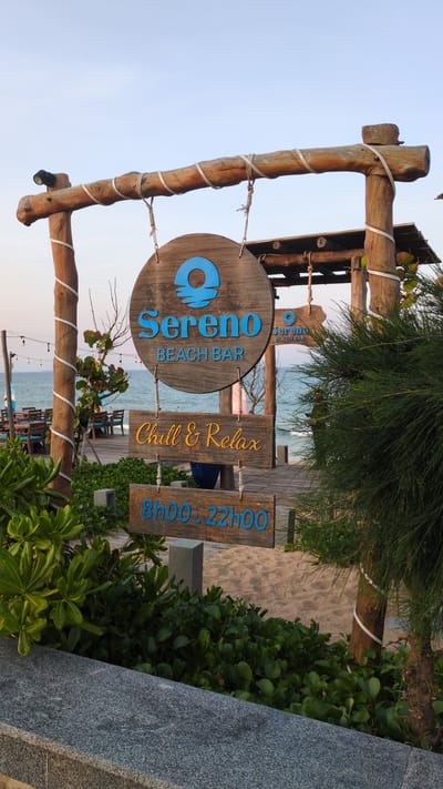 A serene beach bar called "sereno BEACH BAR" is located in Unknown, Vietnam. The bar's name is prominently displayed on a circular wooden sign in blue letters, hung by a rope from a light-colored wooden A-frame wrapped with white tape. Additional signs reading "Chill & Relax" and "8h00 - 22h00" are arranged beside the main sign. The beach bar is accessible via a wooden boardwalk, surrounded by lush green foliage and sand. The ocean is visible in the background.