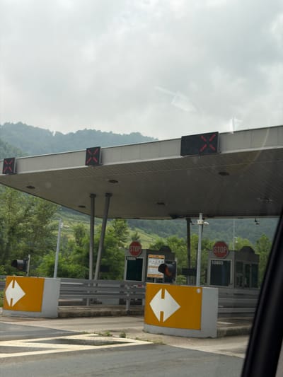 This media file depicts a toll booth on a cloudy day. The scene is viewed from within a vehicle, with the majority of the image focused on the toll plaza infrastructure. The toll booth itself is a modern structure with a gray, textured roof supported by metal poles. Several signs are visible, including red "STOP" signs with white lettering, and black signs with illuminated red "X" symbols, likely indicating lane closures. There are also yellow signs with white arrow symbols, suggesting directions or lane usage. A number, "10805," is displayed on a sign. Traffic lights are also present. In the background, a lush, green hillside with trees is visible, hinting at a rural or mountainous setting. The sky is overcast and white, suggesting a lack of direct sunlight. The overall impression is of a functional toll collection point, likely on a highway or major road in Jehovina, Bosnia and Herzegovina. No people are clearly visible, and no specific emotions are conveyed.