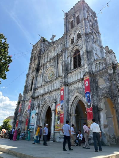 The image shows the exterior of a large, ornate church with a tall bell tower. The church is made of weathered, grey stone and features gothic-style arches, intricate carvings, and a large rose window. Several red banners with religious imagery and text in Vietnamese are hanging on the facade. A cross is visible at the top of the bell tower. In front of the church, a group of people are gathered, some walking, others standing and conversing. They are dressed in casual clothing, including shirts, pants, and dresses. Some are holding umbrellas or bags. The ground in front of the church is a paved walkway. The sky is blue with some scattered white clouds. The location is identified as the church in Hội Tín, Vietnam.