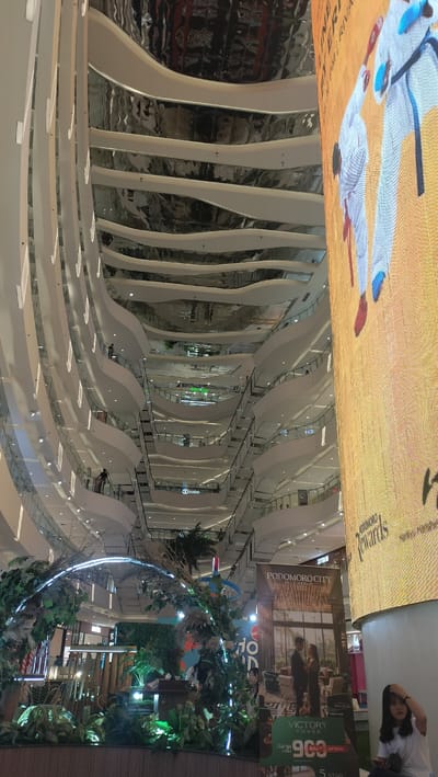 The image captures the interior of a multi-level shopping mall in the City of Medan, Indonesia. The architecture features a distinctive undulating design with multiple curved balconies and escalators visible. A large, illuminated digital screen dominates the right side of the frame, displaying an advertisement. On the left, a decorative archway adorned with greenery and lights is positioned in the foreground. Several individuals are dispersed throughout the mall, engaged in various activities such as walking and standing on the balconies. The overall environment is brightly lit, suggesting it's a public indoor space.