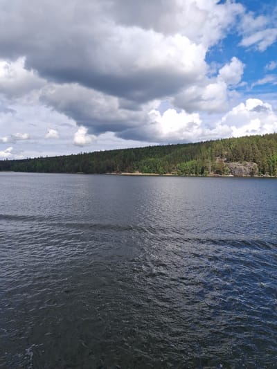 This detailed image captures a natural landscape featuring a large body of water, a forested shoreline, and a dynamic sky. The foreground and midground consist of dark, rippled water with numerous small waves, indicating surface movement. The distant bank is a continuous, dense forest primarily composed of coniferous trees, intermixed with some deciduous foliage, covering low hills and slopes. Approximately in the center-right of the distant shoreline, a prominent dark grey rocky outcrop extends to the water's edge, partially overgrown with trees. To the left of this rock formation, a narrow, light-colored strip, possibly a sandy or gravel beach, is visible along the water's edge. The sky above is partly cloudy, with large, dark grey cumulus clouds dominating the upper left and central portions of the frame. Towards the upper right, patches of vibrant blue sky are visible between brighter white cumulus clouds. There are no individuals, animals, or man-made objects discernible within the image. The scene portrays a tranquil, natural environment characteristic of the Södertälje region in Sweden.