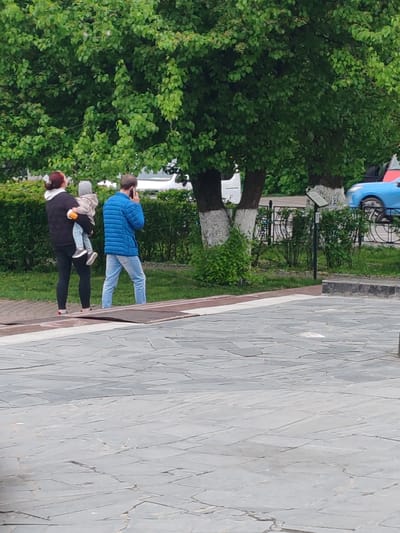 A family – a woman holding a child and a man on a phone call – stands near a sidewalk on gray paving stones. The family stands in front of green bushes and trees with white paint on the lower portions of the trunks. Behind the trees and bushes, a blue car and a white vehicle are visible. A sign is near the bushes and trees. The photo likely was taken in a public area of Tver, Russia, considering the urban features.