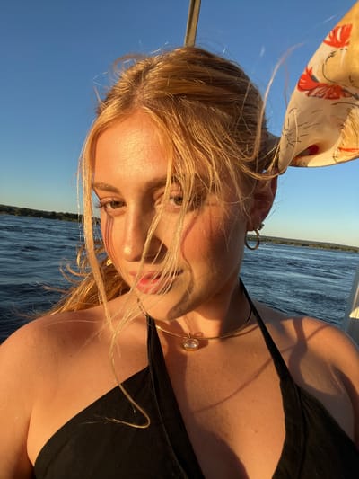 A woman with blonde hair and a headscarf is on a boat. She is wearing a black top and gold jewelry. The background is the Zambezi River at sunset, near Victoria Falls, Zimbabwe.